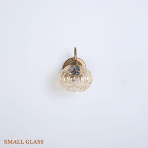 Puede incluir: Una peque&ntilde;a l&aacute;mpara de pared de vidrio con acabado de lat&oacute;n. La l&aacute;mpara tiene una pantalla de vidrio redonda de color &aacute;mbar con detalles acanalados verticales. El texto "SMALL GLASS" est&aacute; en la parte inferior de la imagen.