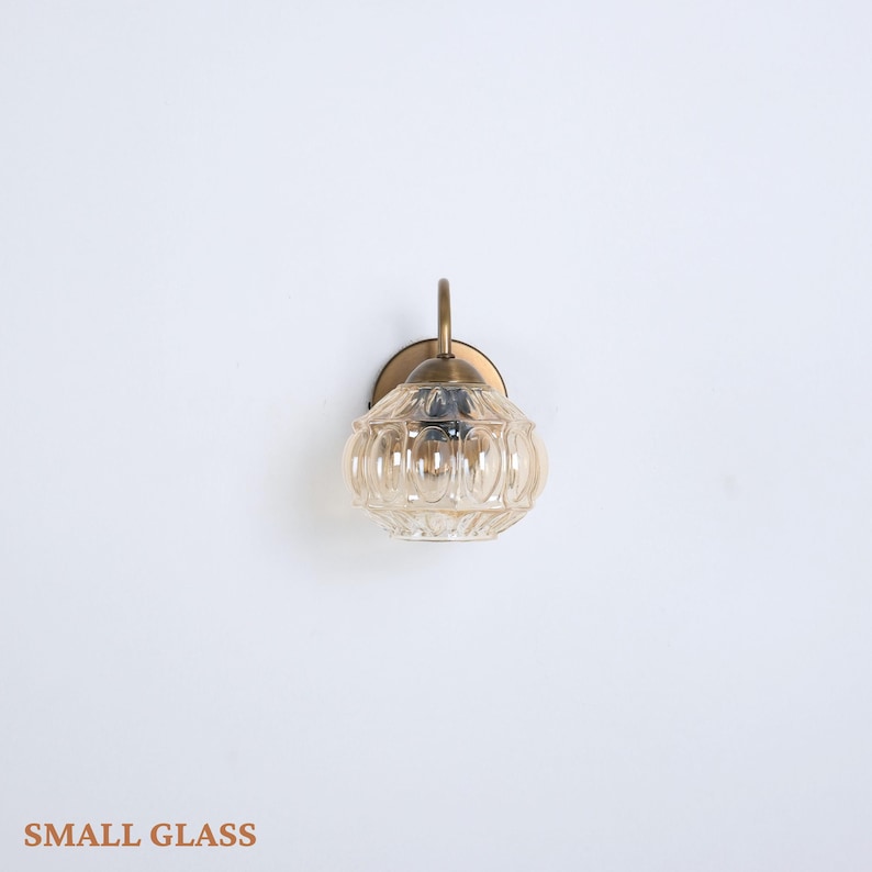 Puede incluir: Una peque&ntilde;a l&aacute;mpara de pared de vidrio con acabado de lat&oacute;n. La l&aacute;mpara tiene una pantalla de vidrio redonda de color &aacute;mbar con detalles acanalados verticales. El texto "SMALL GLASS" est&aacute; en la parte inferior de la imagen.