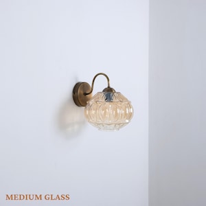 Puede incluir: L&aacute;mpara de pared con una pantalla de cristal color &aacute;mbar con un patr&oacute;n repetido. La l&aacute;mpara tiene un brazo curvo de color lat&oacute;n y una base circular. El texto "MEDIUM GLASS" est&aacute; en la parte inferior.