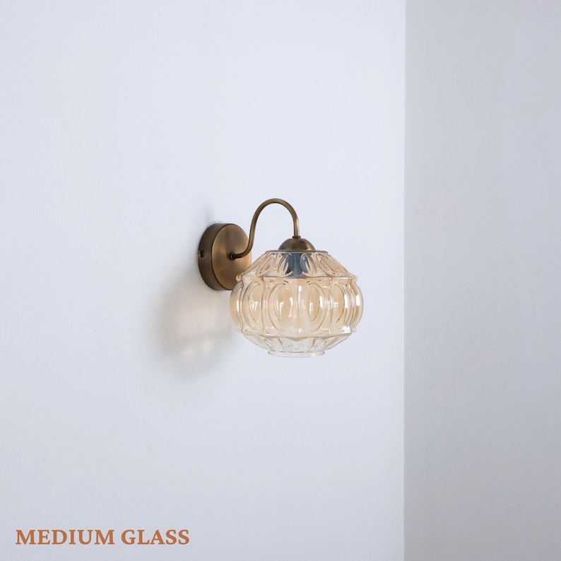 Puede incluir: L&aacute;mpara de pared con una pantalla de cristal color &aacute;mbar con un patr&oacute;n repetido. La l&aacute;mpara tiene un brazo curvo de color lat&oacute;n y una base circular. El texto "MEDIUM GLASS" est&aacute; en la parte inferior.