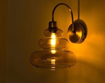 Könnte beinhalten: Eine goldene Wandleuchte mit einem mehrschichtigen, bernsteinfarbenen Glasschirm. Das Licht ist eingeschaltet und beleuchtet den Raum.