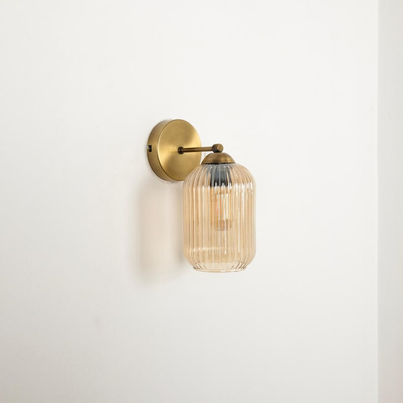 May include: A wall-mounted light fixture with a round, brass-colored base and a cylindrical, ribbed glass shade. The shade is amber-colored, and the fixture has a vintage aesthetic. The light bulb is visible inside the shade.