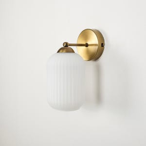 May include: A wall-mounted light fixture with a round, brass-colored base and a cylindrical, ribbed, frosted glass shade. The fixture is attached to the wall with a brass-colored arm. The overall design is minimalist and modern.