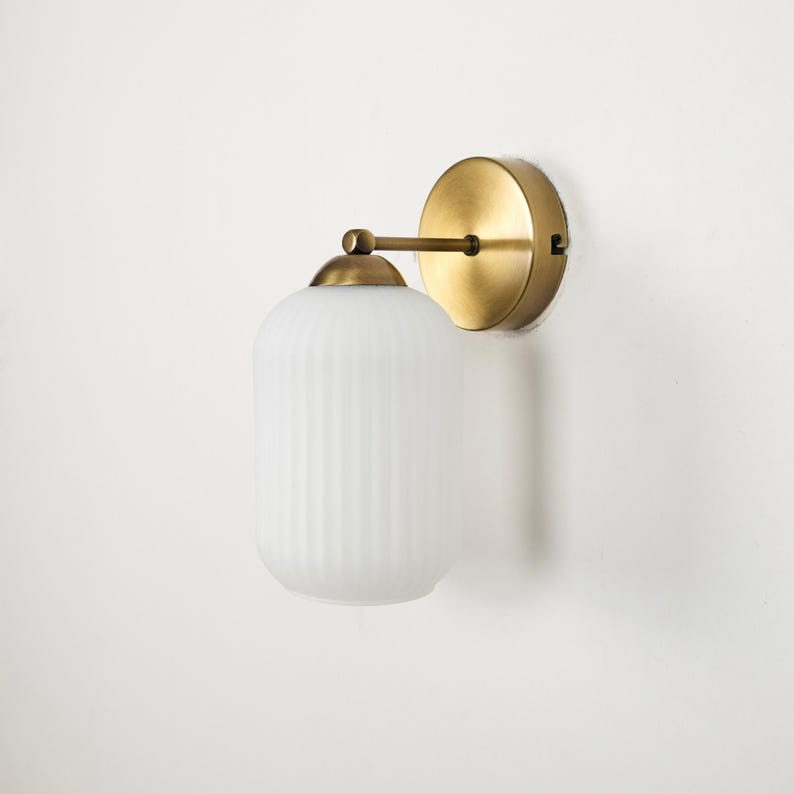 May include: A wall-mounted light fixture with a round, brass-colored base and a cylindrical, ribbed, frosted glass shade. The fixture is attached to the wall with a brass-colored arm. The overall design is minimalist and modern.