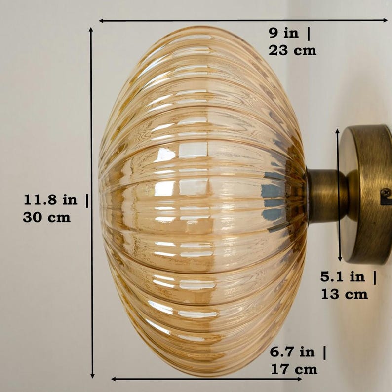 May include: A gold-toned wall sconce with a ribbed amber glass shade. The shade is 9 inches in diameter and 11.8 inches tall. The sconce is 6.7 inches wide and 5.1 inches deep.