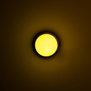 Op de afbeelding: Een ronde plafondlamp met een geel licht dat schijnt door een witte diffuser.