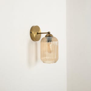 Puede incluir: Una lámpara de pared con acabado de latón. Cuenta con una base redonda y un brazo curvo que sostiene una pantalla de vidrio ámbar acanalado. La bombilla es visible dentro de la pantalla. La lámpara está montada en una pared blanca.