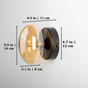 May include: A round amber glass wall sconce with a black metal base. The sconce measures 5.5 inches in diameter and 4.7 inches in height. The base is 3.1 inches in diameter.
