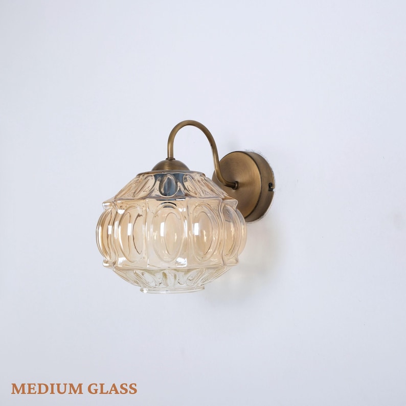 Puede incluir: Aplique de pared con una pantalla de cristal transparente de color &aacute;mbar. La pantalla tiene un patr&oacute;n ovalado y est&aacute; sostenida por un brazo curvo de tono lat&oacute;n y una base circular. El texto "MEDIUM GLASS" est&aacute; en la parte inferior.
