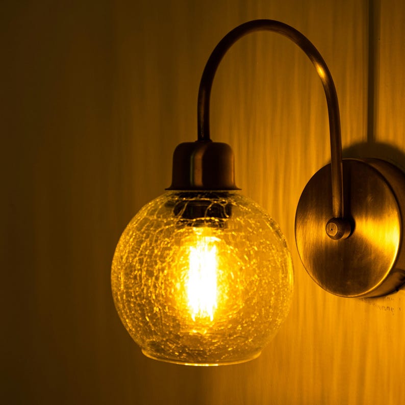 K&ouml;nnte beinhalten: Eine Wandleuchte aus Messing mit einem runden, strukturierten Glasschirm. Das Licht ist an und die Gl&uuml;hbirne leuchtet.