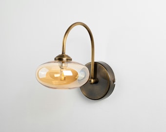Può includere: Una lampada da parete con un braccio curvo color ottone che supporta un paralume rotondo in vetro ambrato. L'apparecchio è fissato a una base circolare scura, creando un'estetica vintage.