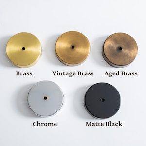 May include: Five round ceiling canopy options in brass, vintage brass, aged brass, chrome, and matte black. Each canopy has a central hole. The names of the finishes are printed below each canopy.