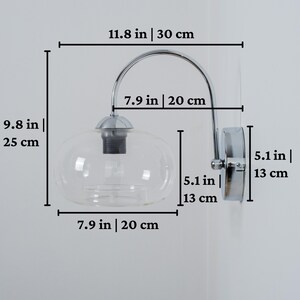 May include: Chrome wall sconce with a clear glass globe shade. The sconce measures 11.8 inches wide, 9.8 inches tall, and 5.1 inches deep.