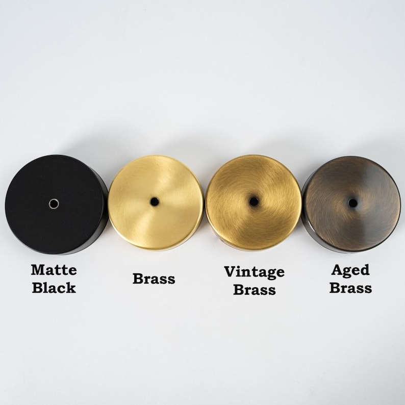 Op de afbeelding: Vier ronde metalen doppen in verschillende afwerkingen: mat zwart, messing, vintage messing en gepatineerd messing. Elke dop heeft een klein gaatje in het midden.