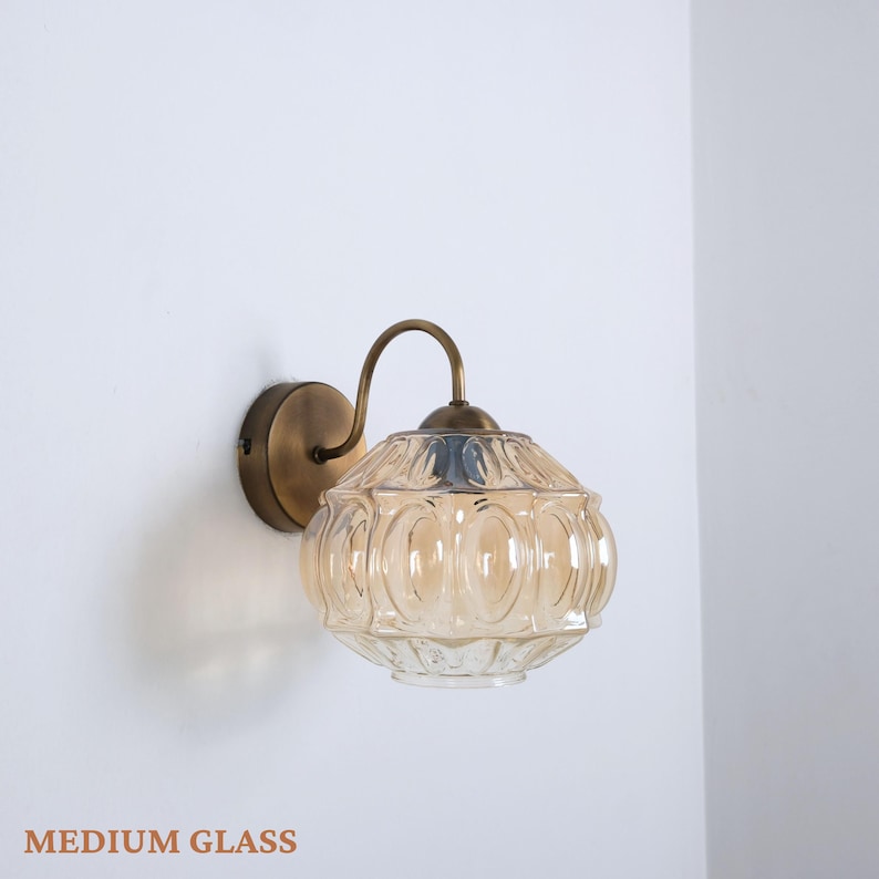 Puede incluir: L&aacute;mpara de pared con una pantalla de vidrio &aacute;mbar de forma redonda y con un dise&ntilde;o texturizado. El brazo y la base son de color lat&oacute;n. El texto "MEDIUM GLASS" se encuentra en la parte inferior.