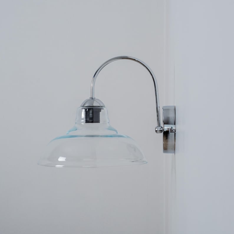 May include: A chrome wall-mounted light fixture with a clear glass shade. The shade is bell-shaped and has a rounded top.