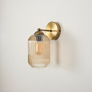May include: A wall-mounted light fixture with a clear, ribbed glass shade in a warm amber color. The fixture has a brass-colored metal base and arm, and a visible lightbulb. The design is simple and elegant.