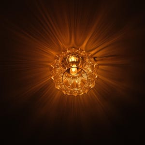 May include: A close-up shot of a vintage-style glass light fixture. The fixture has a decorative, scalloped edge and a central light bulb. The light casts a warm, golden glow, creating a starburst effect against a dark background.