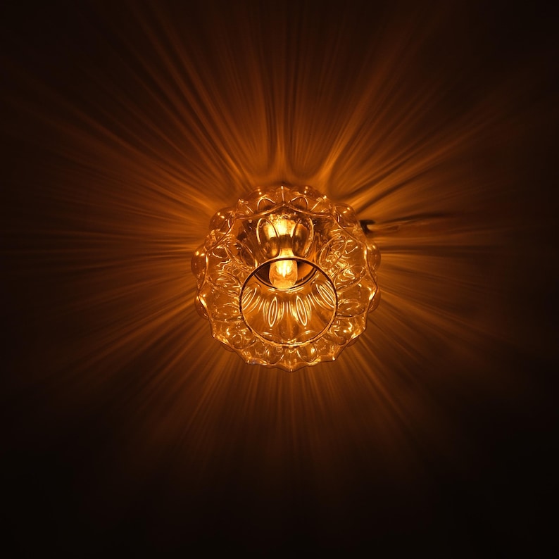 May include: A close-up shot of a vintage-style glass light fixture. The fixture has a decorative, scalloped edge and a central light bulb. The light casts a warm, golden glow, creating a starburst effect against a dark background.