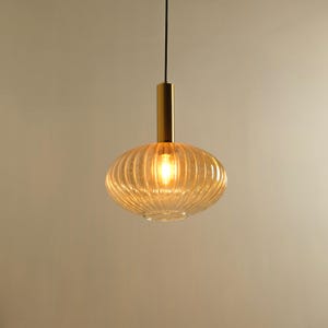 Peut inclure: Une suspension lumineuse dotée d'un abat-jour en verre nervuré teinté ambre, de forme arrondie et aplatie. L'abat-jour est suspendu par un fin cordon noir fixé à une monture cylindrique en métal doré brossé au sommet central.