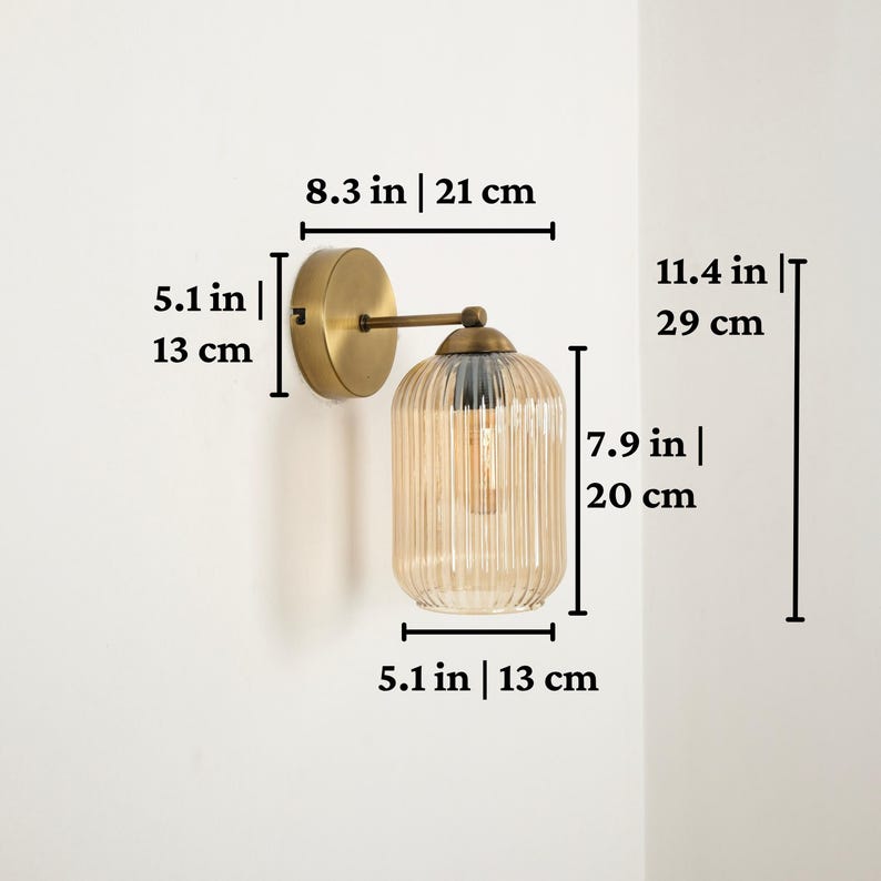 May include: A wall sconce with a gold-colored metal base and arm, supporting a ribbed, amber-colored glass shade. The sconce is 11.4 inches (29 cm) tall, 8.3 inches (21 cm) wide, and 5.1 inches (13 cm) deep. The shade is 7.9 inches (20 cm) tall and 5.1 inches (13 cm) wide.
