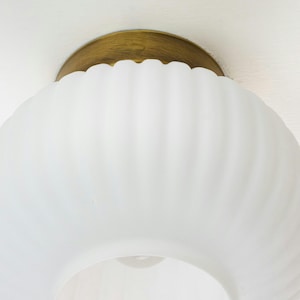 Op de afbeelding: Een witte, gematteerde glazen plafondlamp met een gouden metalen basis. De glazen kap heeft een geribbelde textuur.