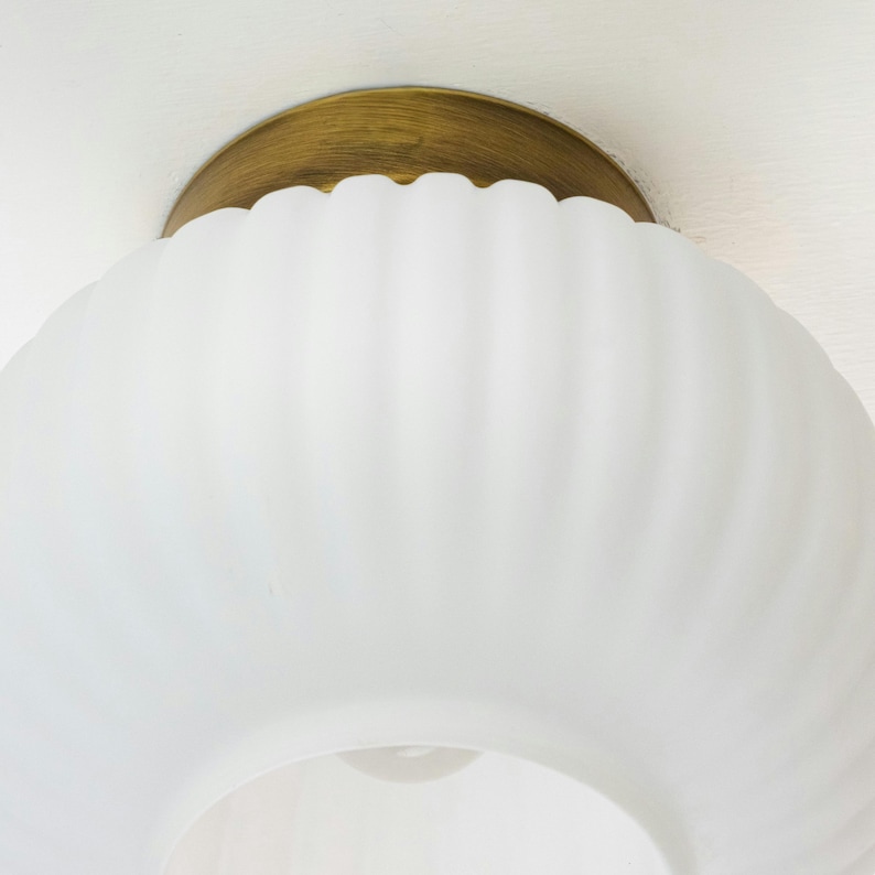 Op de afbeelding: Een witte, gematteerde glazen plafondlamp met een gouden metalen basis. De glazen kap heeft een geribbelde textuur.