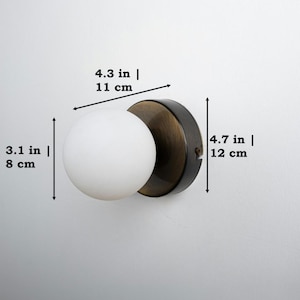 Op de afbeelding: Een zwarte en messing wandlamp met een witte, matte glazen bol. De lamp is 12 cm hoog, 11 cm breed en 8 cm diep.