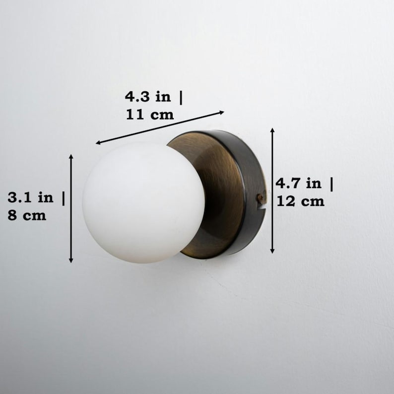 Op de afbeelding: Een zwarte en messing wandlamp met een witte, matte glazen bol. De lamp is 12 cm hoog, 11 cm breed en 8 cm diep.
