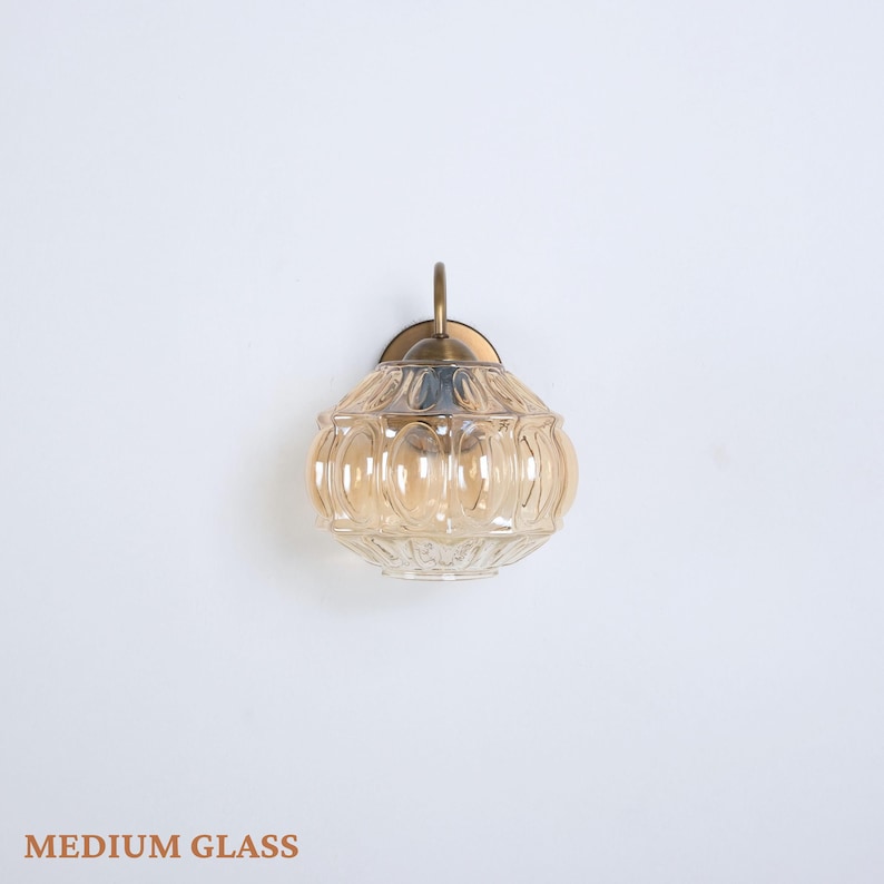 Puede incluir: Aplique de pared de cristal &aacute;mbar de tama&ntilde;o mediano con un dise&ntilde;o decorativo. Tiene una placa de montaje de metal color lat&oacute;n y un brazo curvo. El texto "MEDIUM GLASS" est&aacute; en la parte inferior de la imagen.