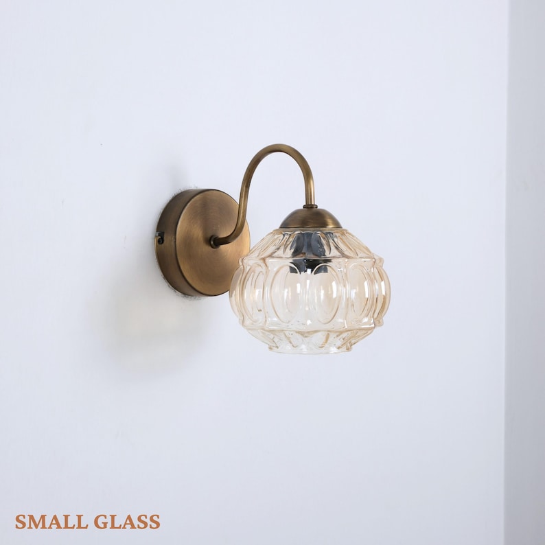 Puede incluir: Una l&aacute;mpara de pared con acabado de lat&oacute;n. La l&aacute;mpara presenta un brazo curvo que sostiene una pantalla redonda de vidrio &aacute;mbar con un dise&ntilde;o estampado. La base es circular y tambi&eacute;n de lat&oacute;n. El texto "SMALL GLASS" est&aacute; en la parte inferior.