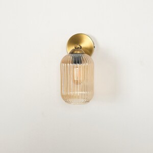 May include: A wall-mounted light fixture with a gold-colored circular base and a ribbed, amber-colored glass shade. The light bulb is visible inside the shade. The fixture is mounted on a white wall.