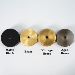 Op de afbeelding: Vier ronde metalen doppen in verschillende afwerkingen: mat zwart, messing, vintage messing en gepatineerd messing. Elke dop heeft een klein gaatje in het midden.