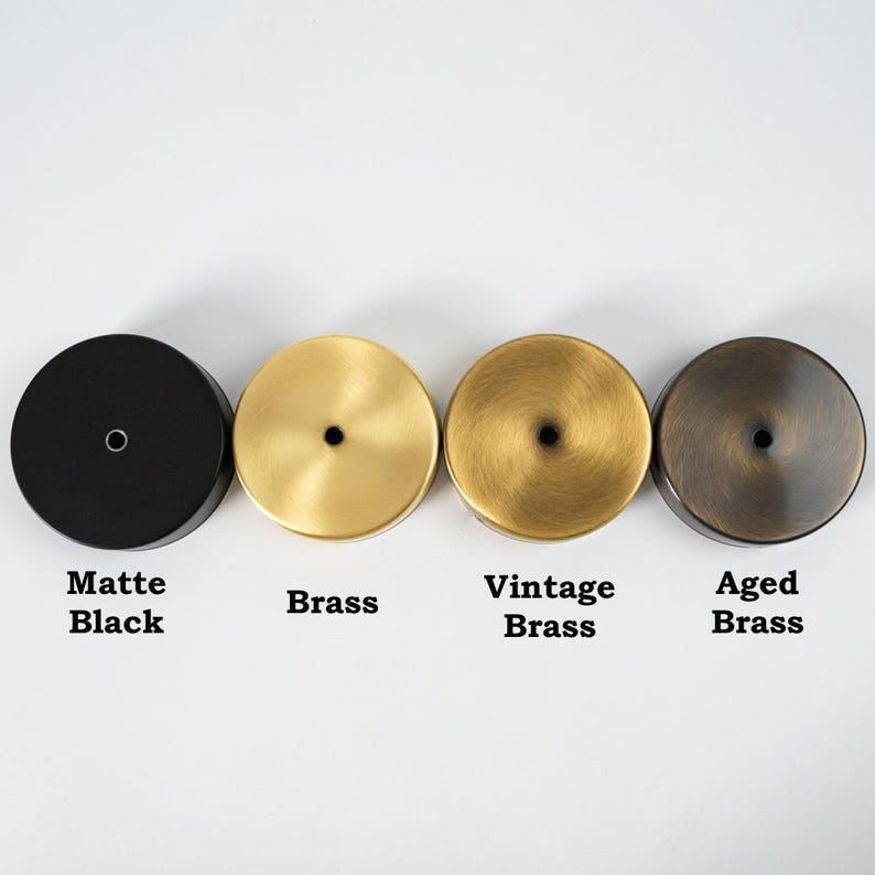 Op de afbeelding: Vier ronde metalen doppen in verschillende afwerkingen: mat zwart, messing, vintage messing en gepatineerd messing. Elke dop heeft een klein gaatje in het midden.