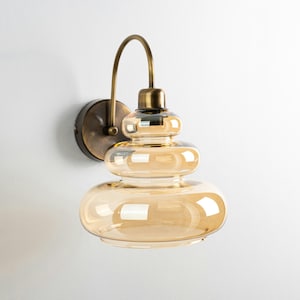 Könnte beinhalten: Eine Wandleuchte aus Messing mit einem mehrschichtigen, bernsteinfarbenen Glasschirm. Die Leuchte hat einen gebogenen Arm und einen runden Sockel.