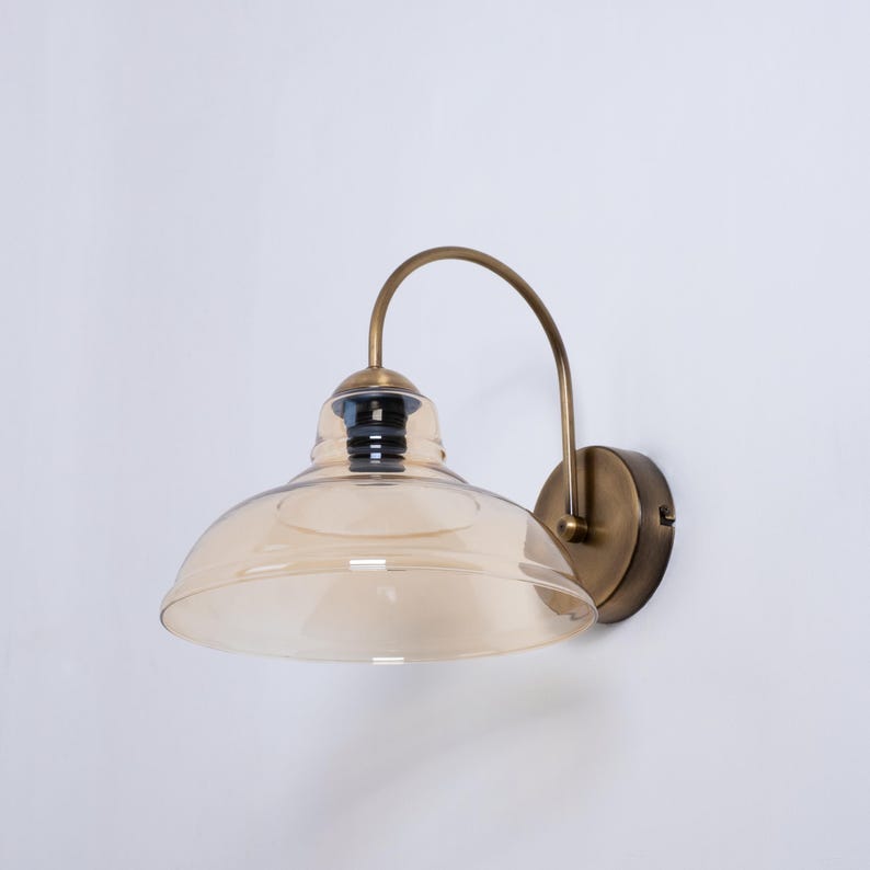 May include: A brass wall sconce with a curved arm and a amber glass shade. The sconce has a simple design and would be a great addition to any home.
