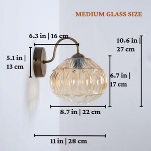 Puede incluir: Una l&aacute;mpara de pared con una pantalla de cristal de color &aacute;mbar. El accesorio tiene una base y un brazo de metal de color bronce. La pantalla mide 17 cm de alto y 22 cm de ancho. La longitud total es de 28 cm. El texto de la imagen dice "MEDIUM GLASS SIZE".
