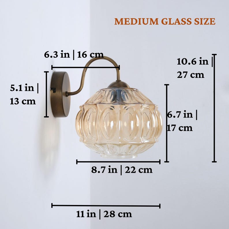 Puede incluir: Una l&aacute;mpara de pared con una pantalla de cristal de color &aacute;mbar. El accesorio tiene una base y un brazo de metal de color bronce. La pantalla mide 17 cm de alto y 22 cm de ancho. La longitud total es de 28 cm. El texto de la imagen dice "MEDIUM GLASS SIZE".