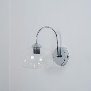 May include: A chrome wall sconce with a clear glass shade. The sconce has a curved arm and a round base.