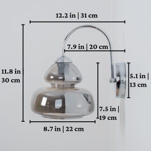 Pu&ograve; includere: Un'applique da parete cromata con un paralume in vetro fum&egrave; a due livelli. L'applique misura 31 cm di altezza, 20 cm di larghezza e 19 cm di profondit&agrave;.