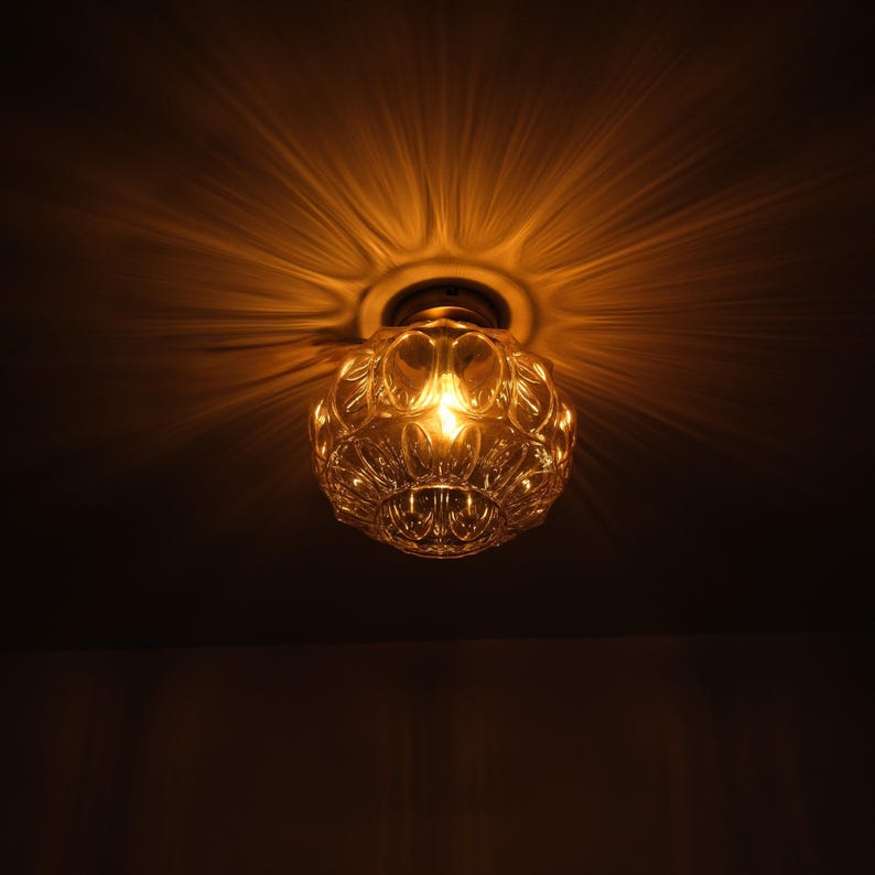 May include: A vintage-style ceiling light fixture with a clear, patterned glass shade. The light is on, casting a warm, golden glow and radiating light patterns on the ceiling. The fixture is mounted on a white ceiling.