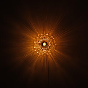 May include: A decorative, ornate light fixture with a central bulb, radiating warm light. The fixture has a detailed, circular design, casting starburst-like shadows against a dark background. The overall effect is a warm, inviting glow.