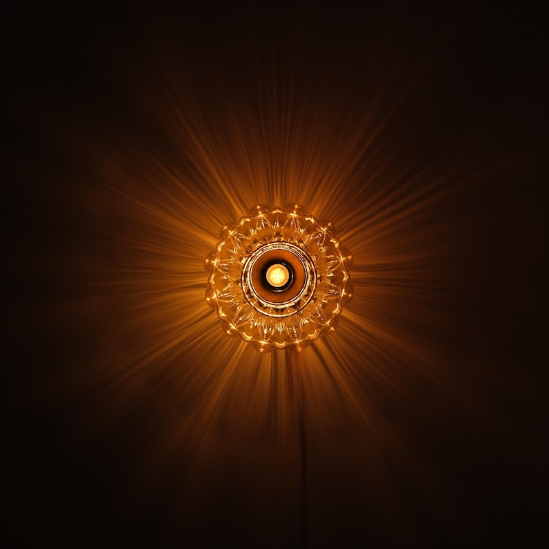 May include: A decorative, ornate light fixture with a central bulb, radiating warm light. The fixture has a detailed, circular design, casting starburst-like shadows against a dark background. The overall effect is a warm, inviting glow.