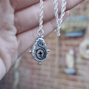 May include: A silver chain necklace with a teardrop-shaped pendant. The pendant has a clear glass or resin insert with a black floral design.