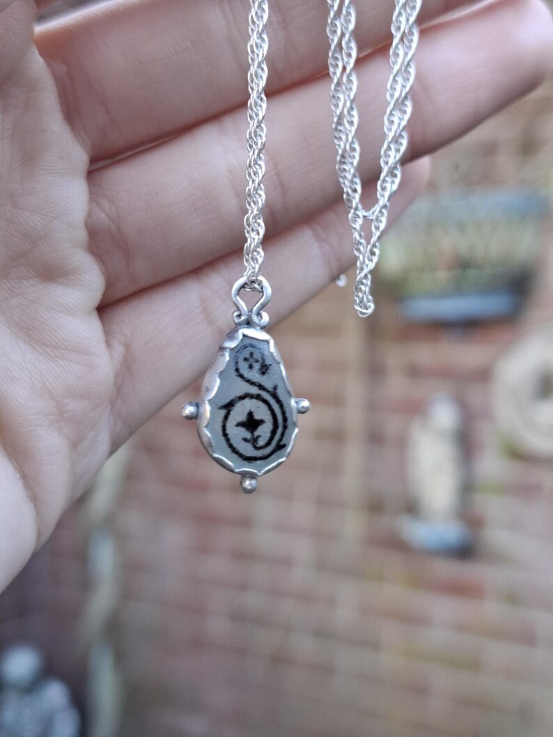 May include: A silver chain necklace with a teardrop-shaped pendant. The pendant has a clear glass or resin insert with a black floral design.