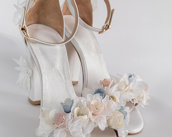 May include: A pair of white high-heeled sandals featuring an ankle strap with a gold-toned buckle and a block heel. The front strap and the back of the heel are decorated with layered fabric flowers in shades of white, pale pink, and light blue.