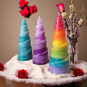Könnte beinhalten: Drei bunte Spiralkerzen mit einem Regenbogenmuster. Die Kerzen stehen auf einer weißen Oberfläche mit einigen roten Rosen.