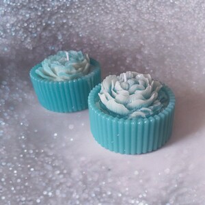 Könnte beinhalten: Zwei tealblaue Kerzenhalter mit weißen Blumenmustern. Die Kerzen sind rosenförmig.