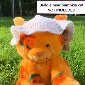 Puede incluir: Un oso de peluche naranja con detalles de calabaza, usando un sombrero blanco de ganchillo. El oso tiene pelaje naranja con parches en forma de calabaza y detalles verdes. El texto "Build a bear pumpkin cat NOT INCLUDED" es visible.