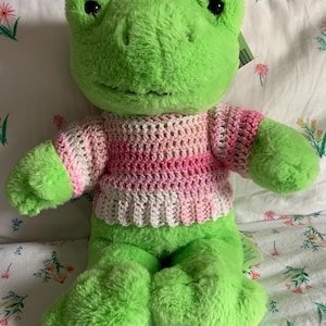 Puede incluir: Un juguete de peluche verde con forma de rana, con ojos negros y un suéter de ganchillo rosa y blanco. La rana está sobre una colcha con estampado floral. El juguete tiene patas grandes y una expresión amigable.